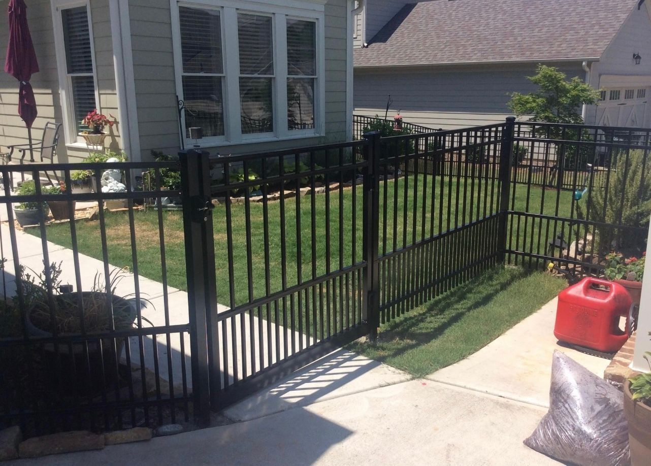 Black metal fence with gate in front of a light-colored house with windows; a red gas can is visible.