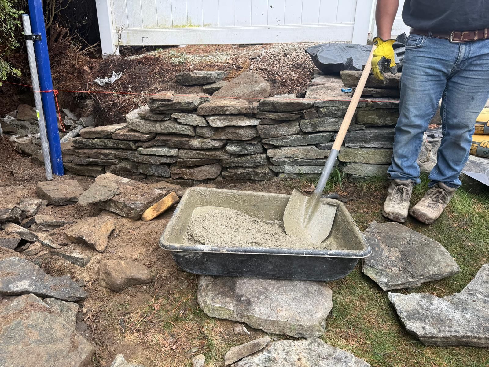 Worker shoveling mortar into a wheelbarrow beside a stacked stone wall and patio renovation area
