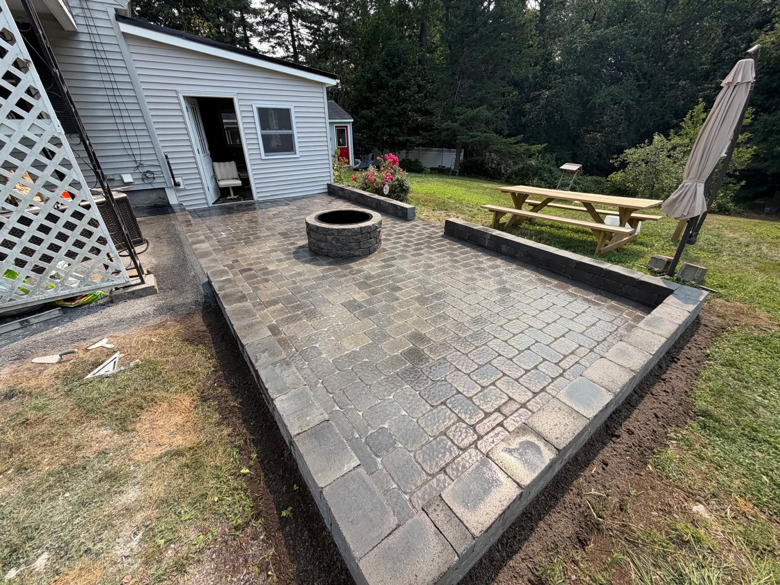 Stone patio beside gray shed with fire pit, picnic table, and umbrella in a backyard
