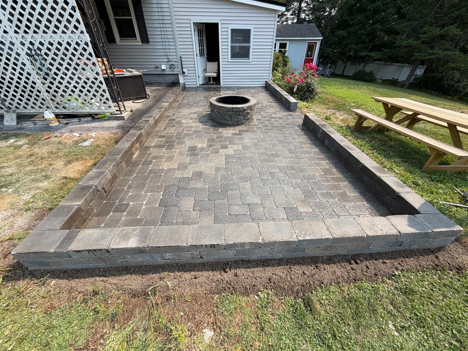 Paver patio with a circular fire pit, bordered by dark edging beside a house and lawn.