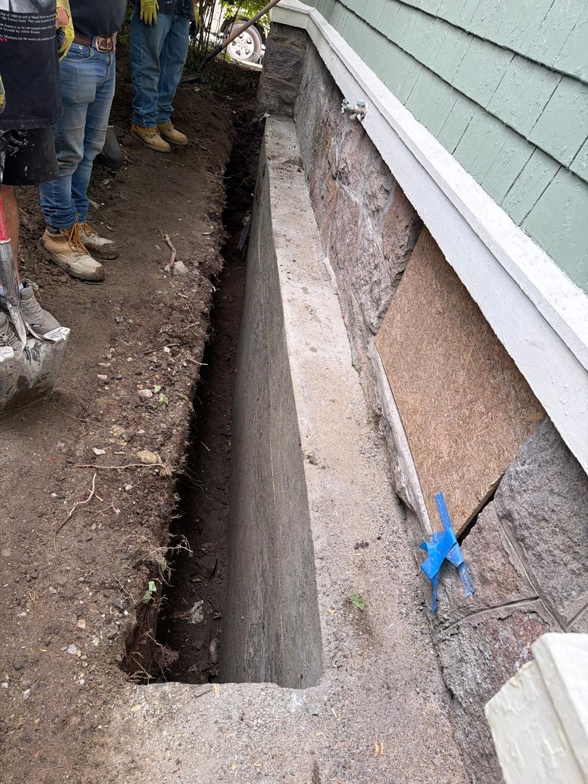 Narrow trench dug beside a house foundation, with workers' boots visible on the left.