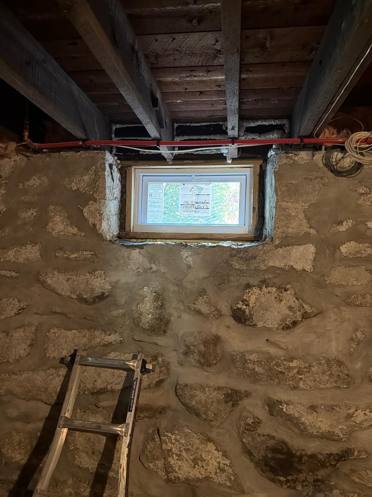 Small basement window in a stone wall beneath exposed ceiling beams, with a ladder leaning below.