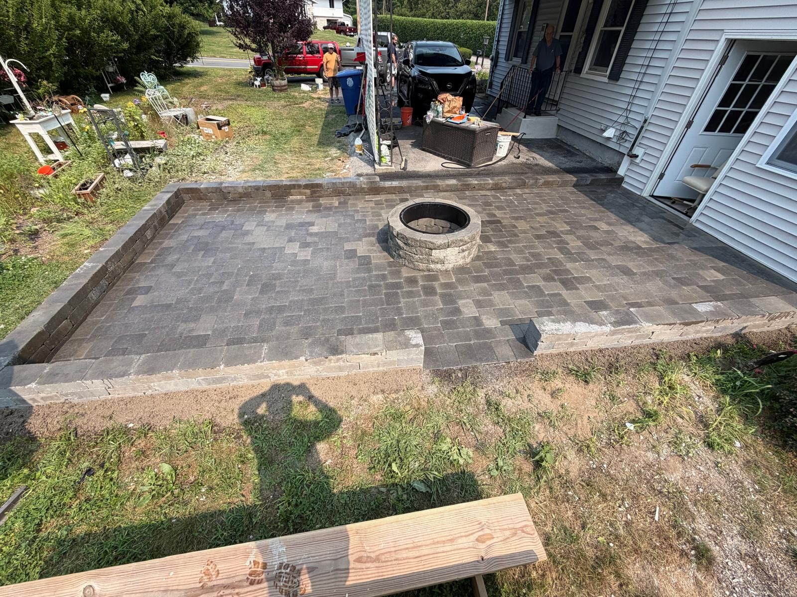 Backyard stone patio with a central fire pit and workers near a house