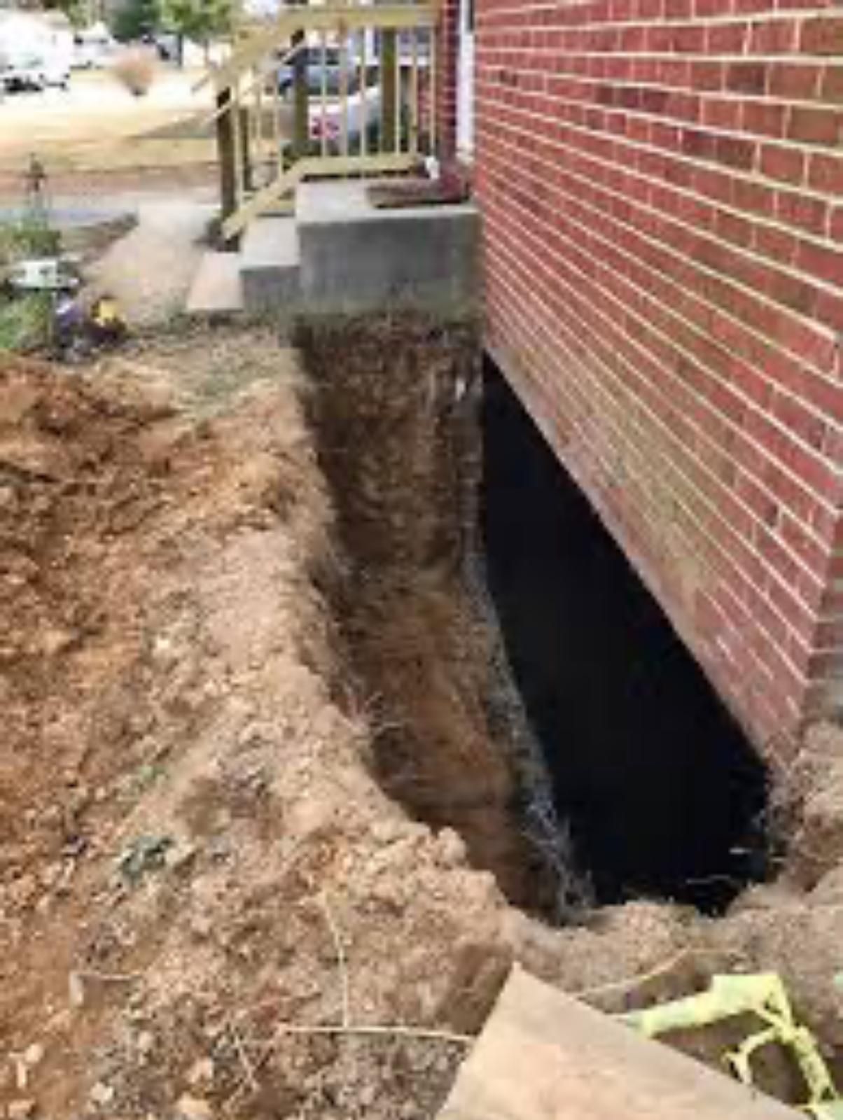 Deep trench dug beside a brick house foundation, exposing a basement opening and soil excavation.