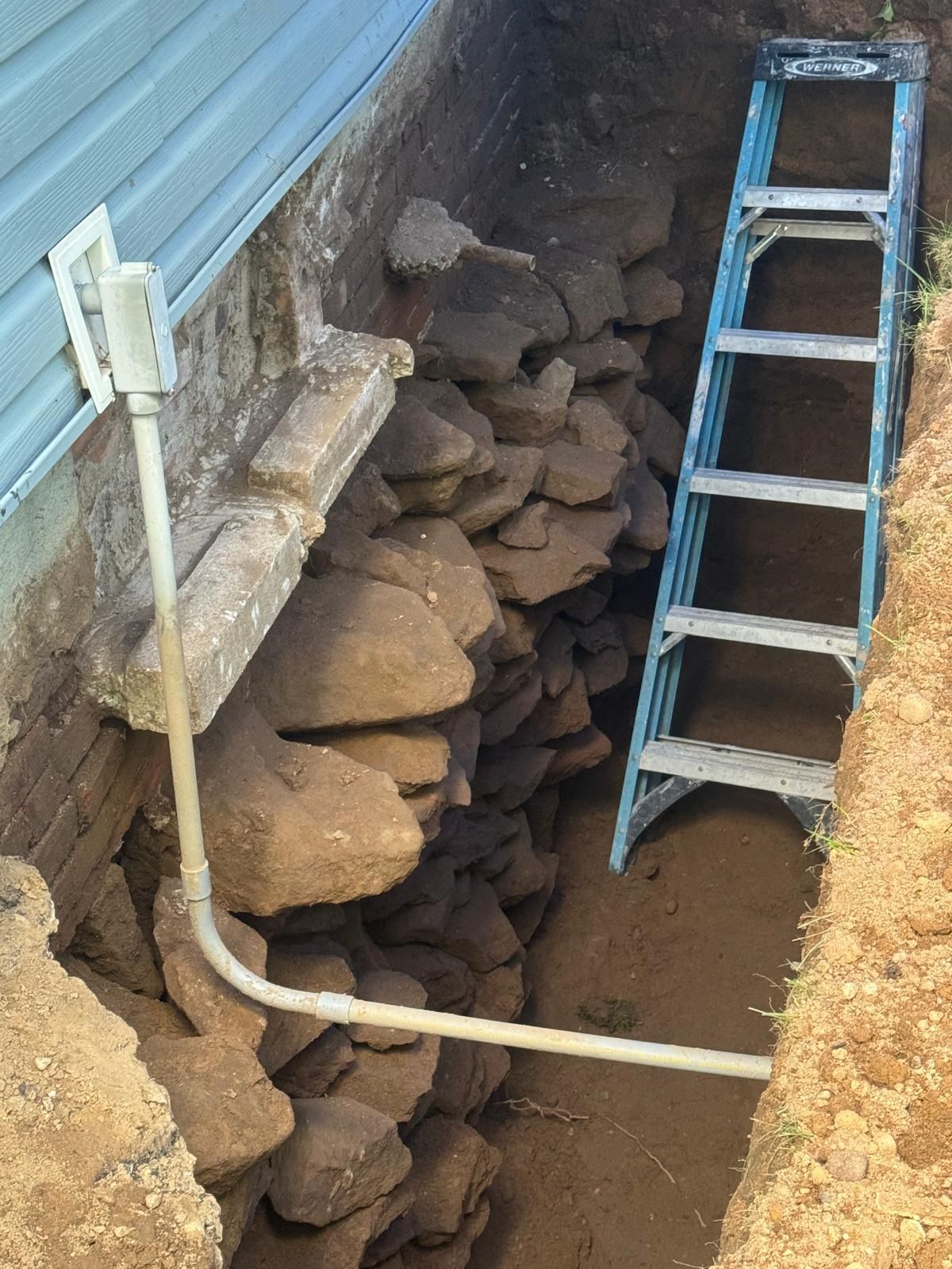 Rock wall collapse beside house foundation with a ladder leaning in a narrow trench