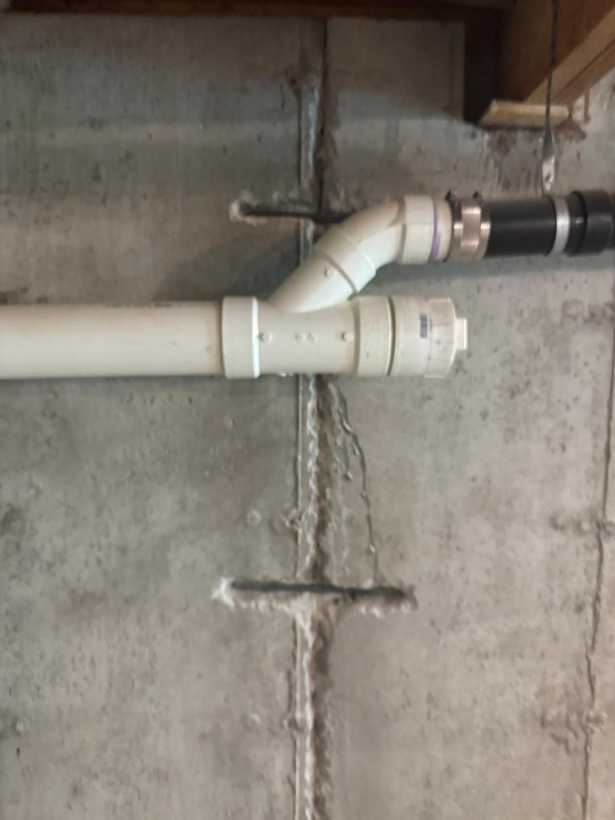 White PVC pipes and fittings running along a concrete wall, joined with a diagonal elbow connection.
