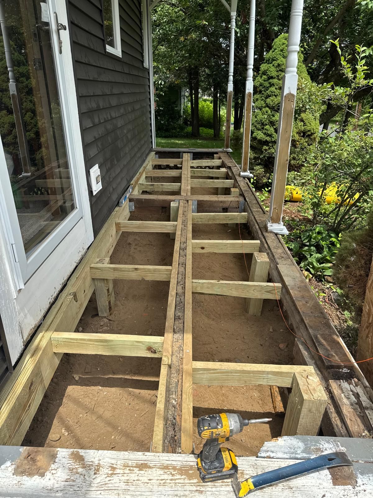 Wooden deck frame under construction beside a house, with tools on the beams and a garden nearby.