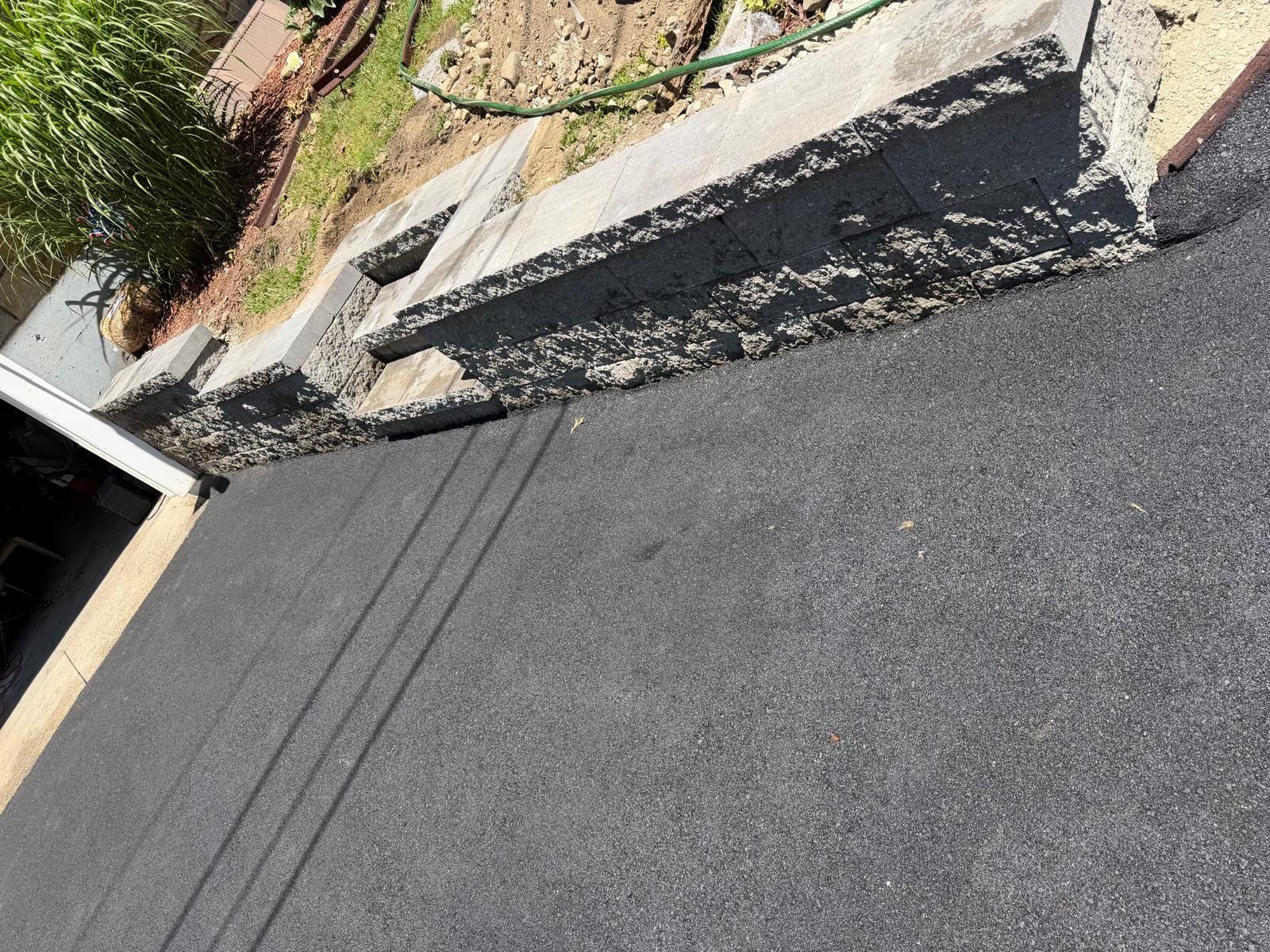 Driveway with freshly paved asphalt beside a concrete retaining wall and garden edging.
