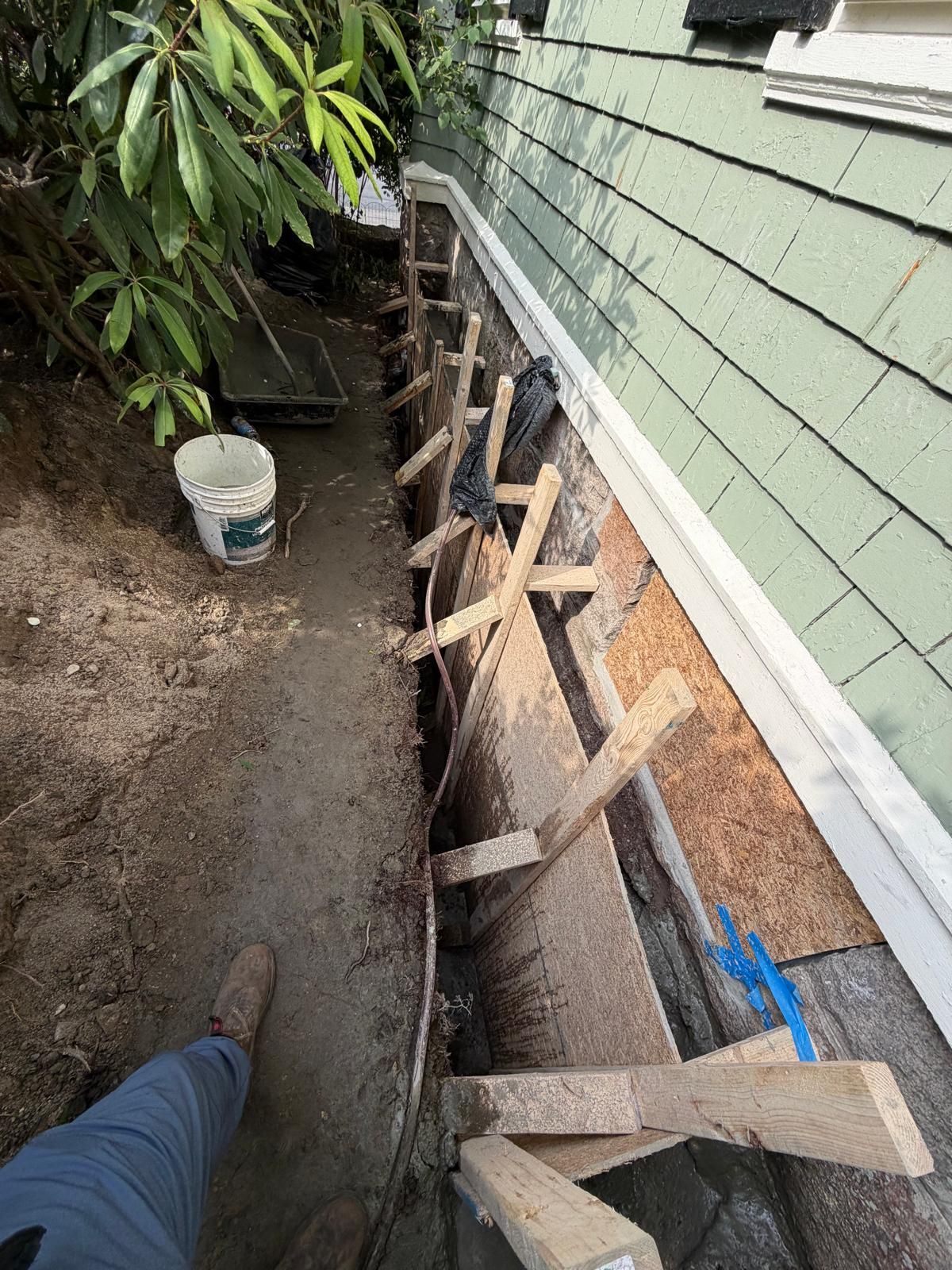 House exterior with exposed framing and trench beside green siding, with a bucket and tools nearby
