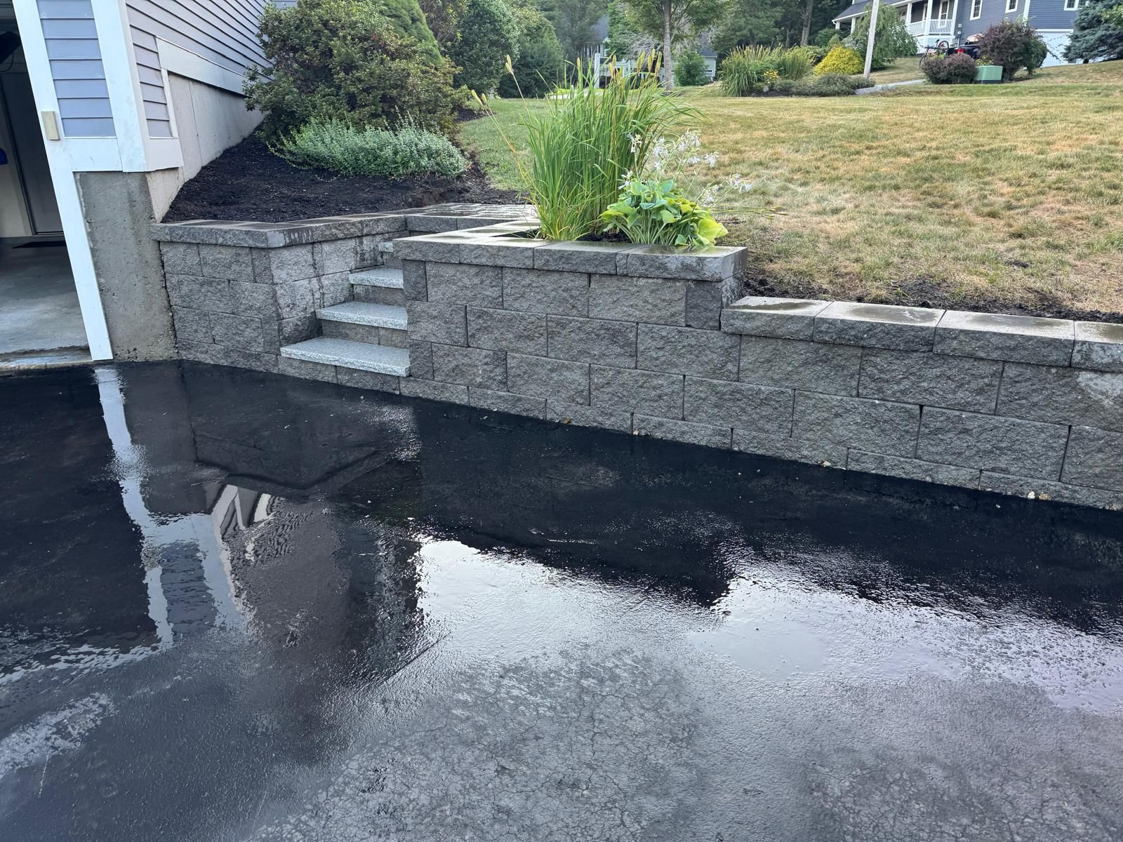Wet driveway beside stone retaining wall and steps leading to a raised yard.