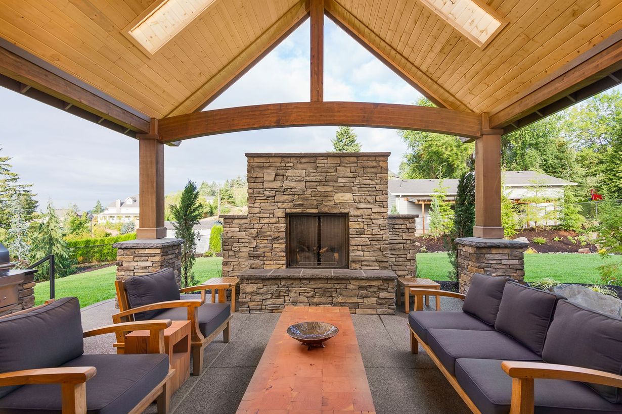 Covered outdoor patio with stone fireplace, wooden beams, and gray seating around a low table.