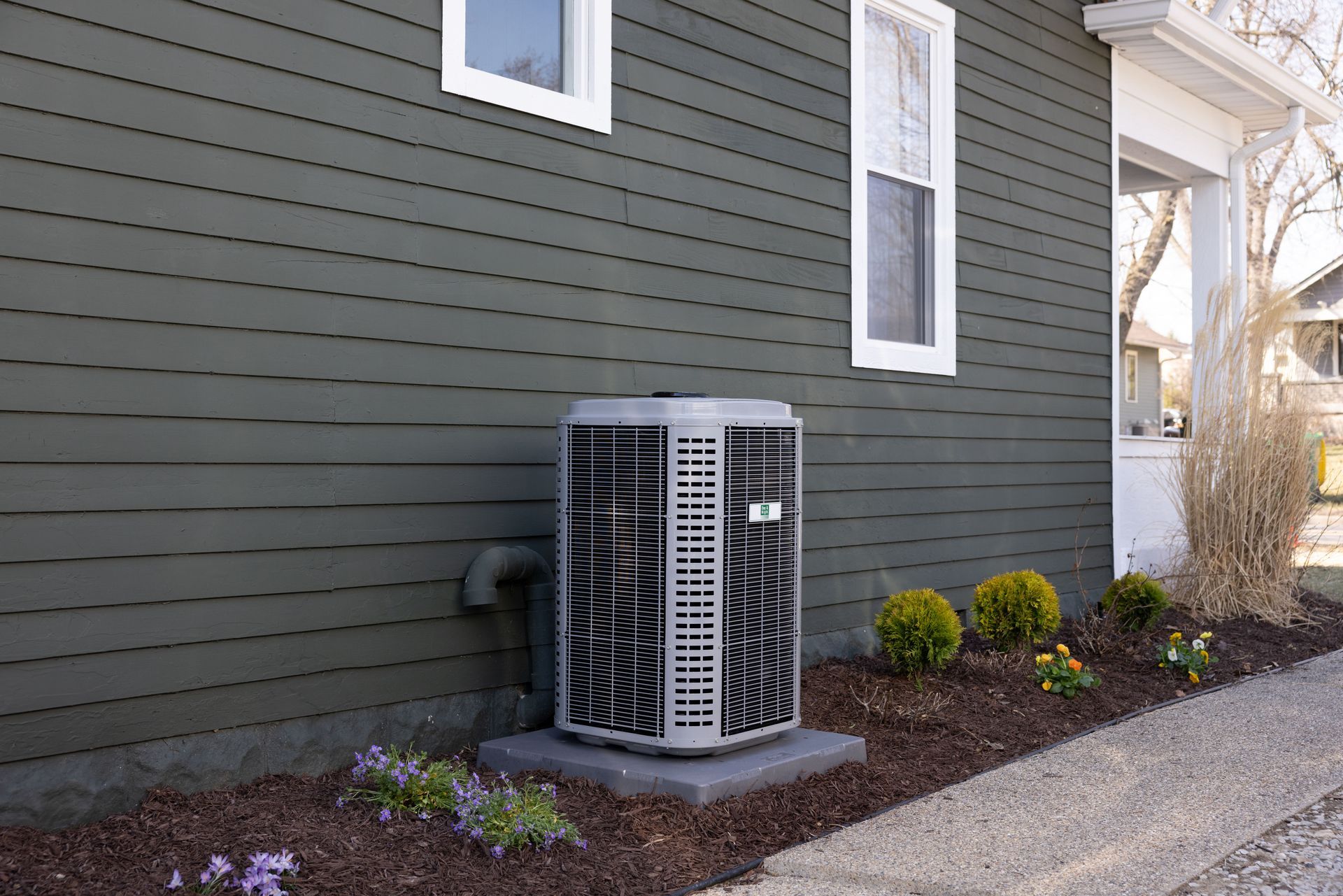 Outdoor HVAC unit beside a dark green house wall and window, next to a gravel path and shrubs