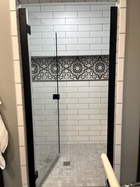 A walk-in shower with a glass door, framed in black, featuring white subway tile walls and a decorative patterned border.