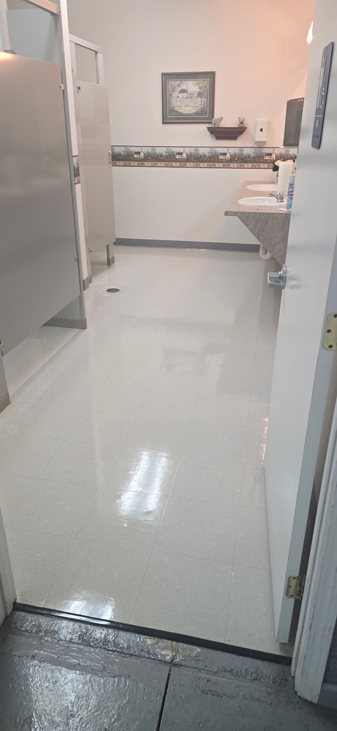 A clean, brightly lit public restroom with light-colored flooring, stall doors on the left, and a sink area at the back.