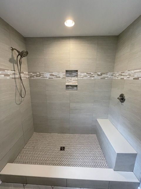 A walk-in shower with gray tile walls, a herringbone patterned floor, a built-in bench, and a handheld shower head.