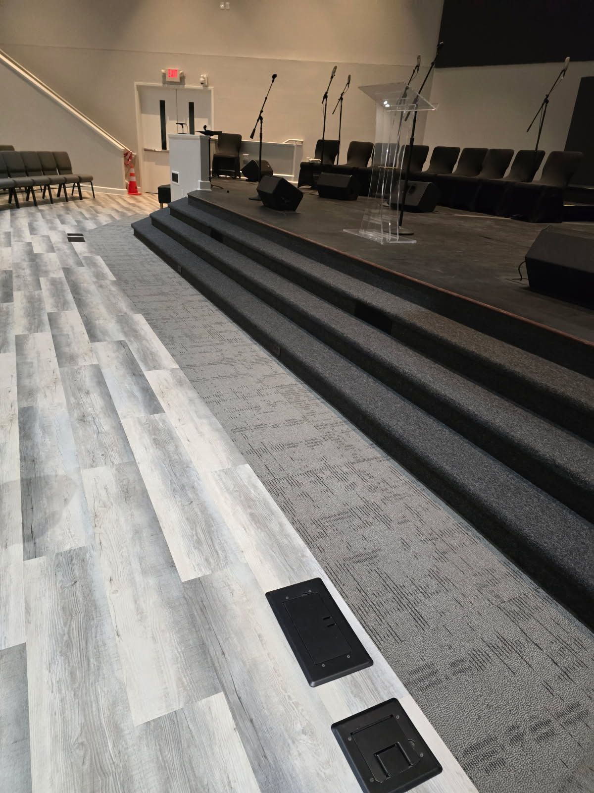 A view of a church stage with steps, a podium, and chairs, featuring light-colored flooring transition to gray carpet.