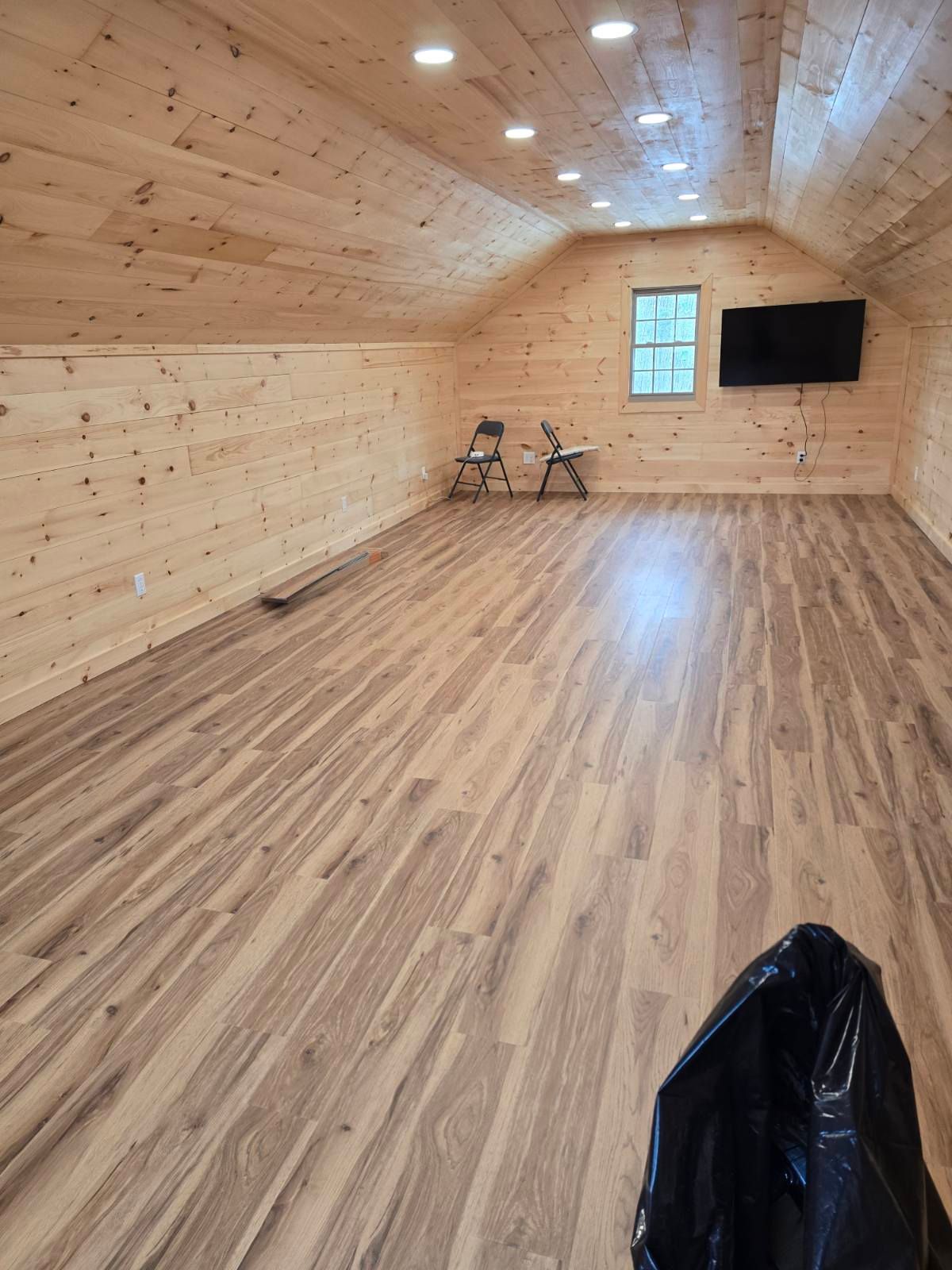 An empty, spacious attic room with light wood walls and flooring, recessed lighting, two chairs, and a wall-mounted TV.
