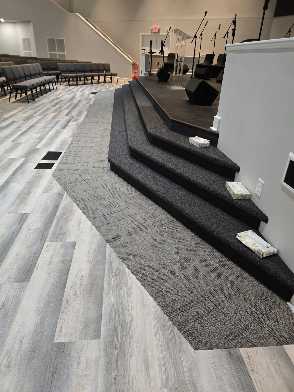 An interior shot of a church stage with steps covered in dark gray carpet, bordered by light wood-look floor planks.