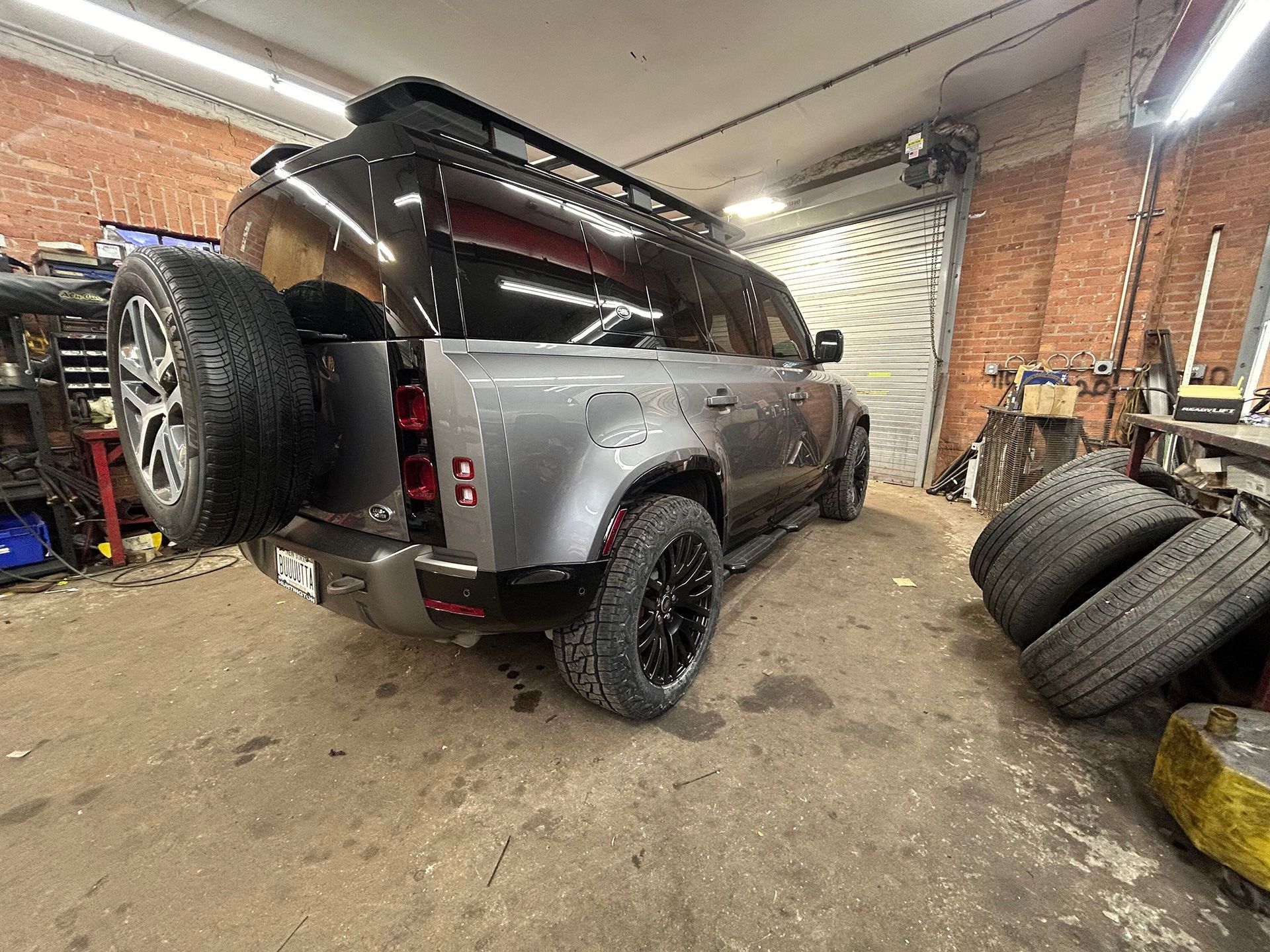 A gray Land Rover Defender is parked in a garage next to a pile of tires