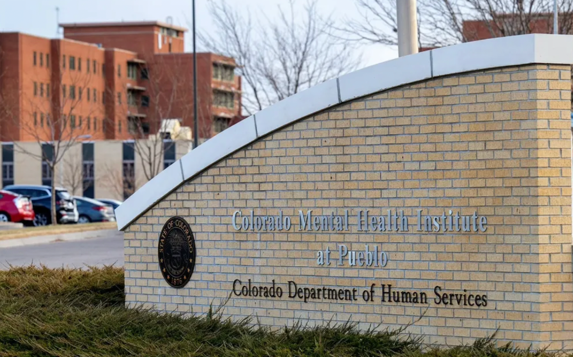 Sign for Colorado Mental Health Institute at Pueblo. Brick wall with text, building in background.
