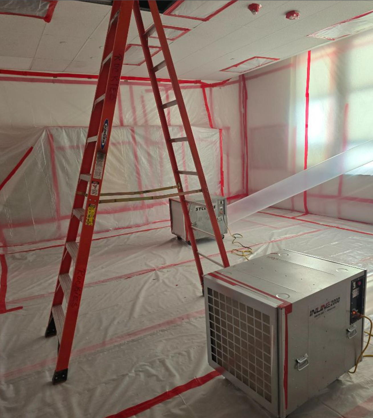 Room enclosed in plastic sheeting, red tape, and a ladder; two air purification units sit on the floor.