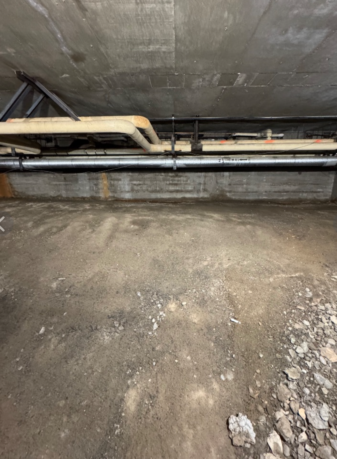 Underground room with pipes along the concrete ceiling and wall. The floor is dirty and dusty.