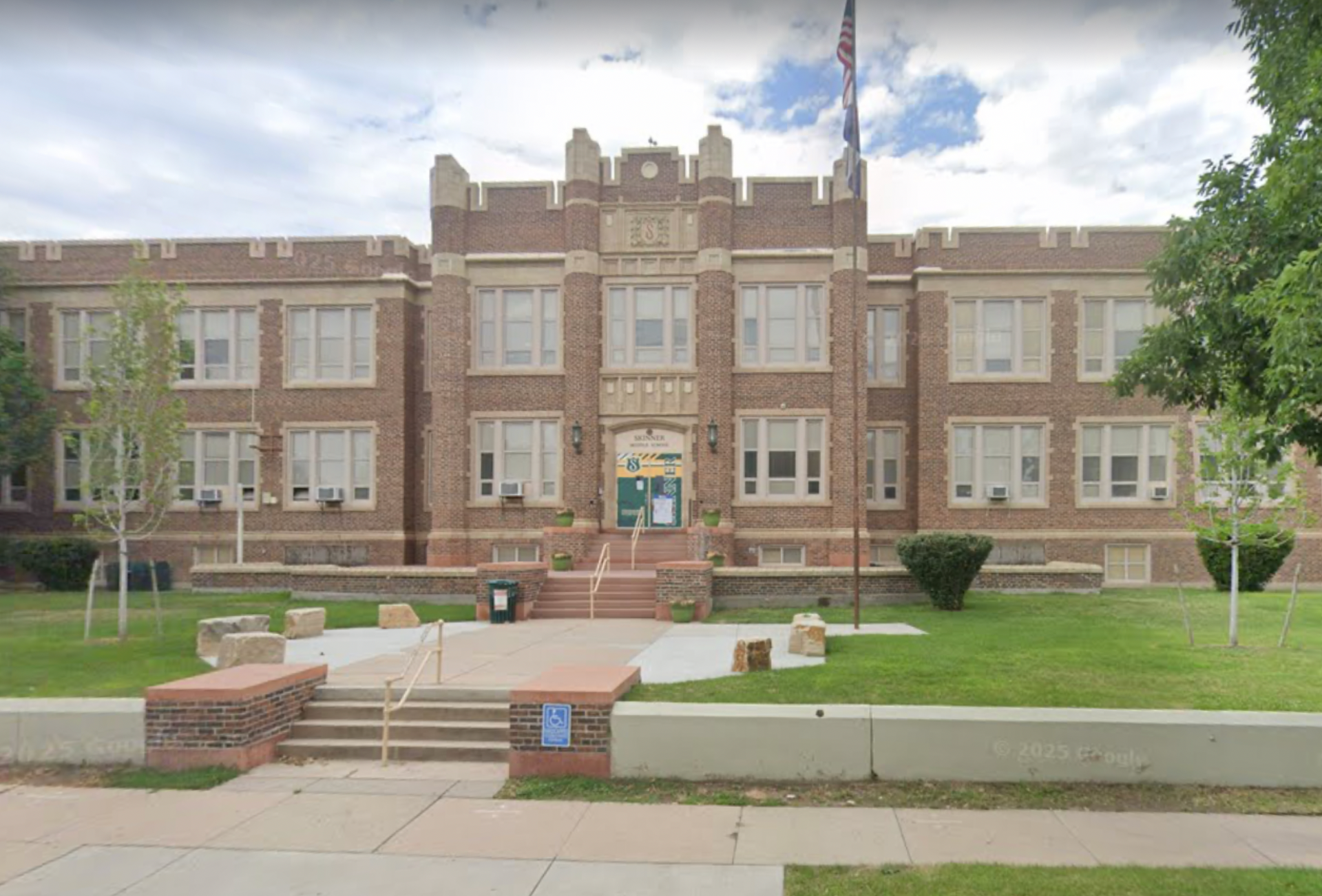 Brick school building with steps, lawn, flag, and cloudy sky.