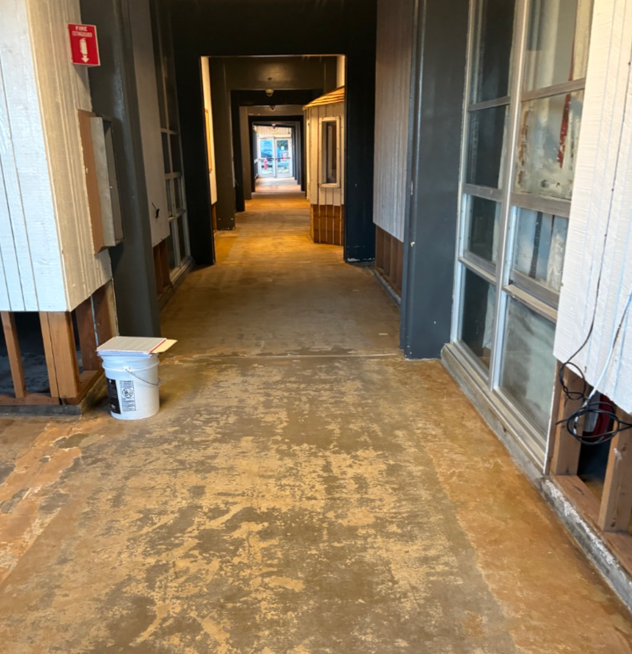 Hallway under renovation with exposed subfloor, white walls, and a fire alarm.