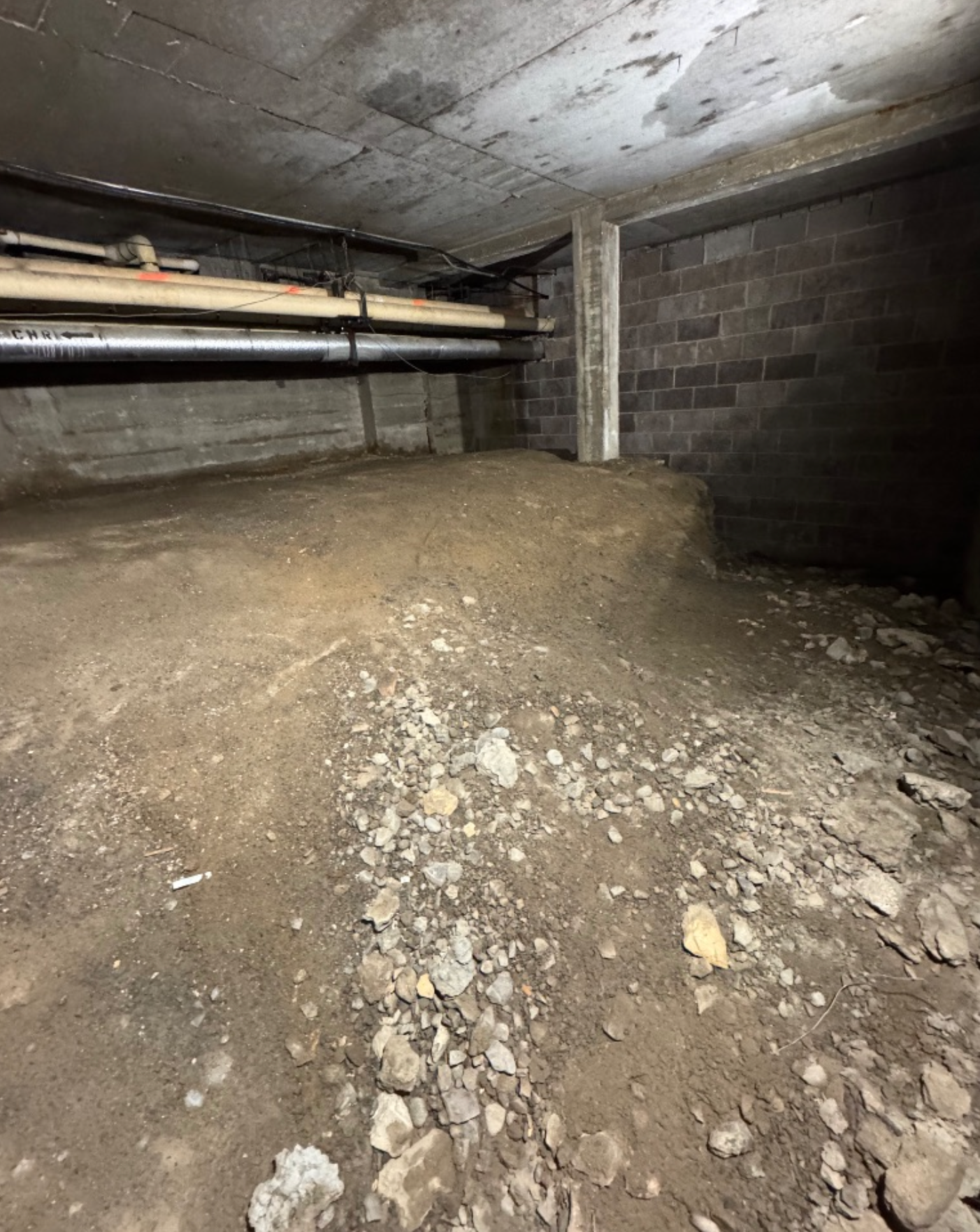 Dirt floor and cinder block walls in a dimly lit basement. Pipes run overhead.