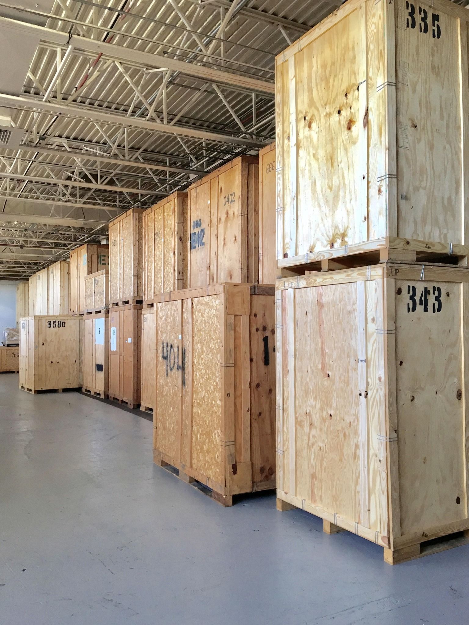 Wooden storage crates stacked in a warehouse with a high ceiling. Crates have labels and numbers.
