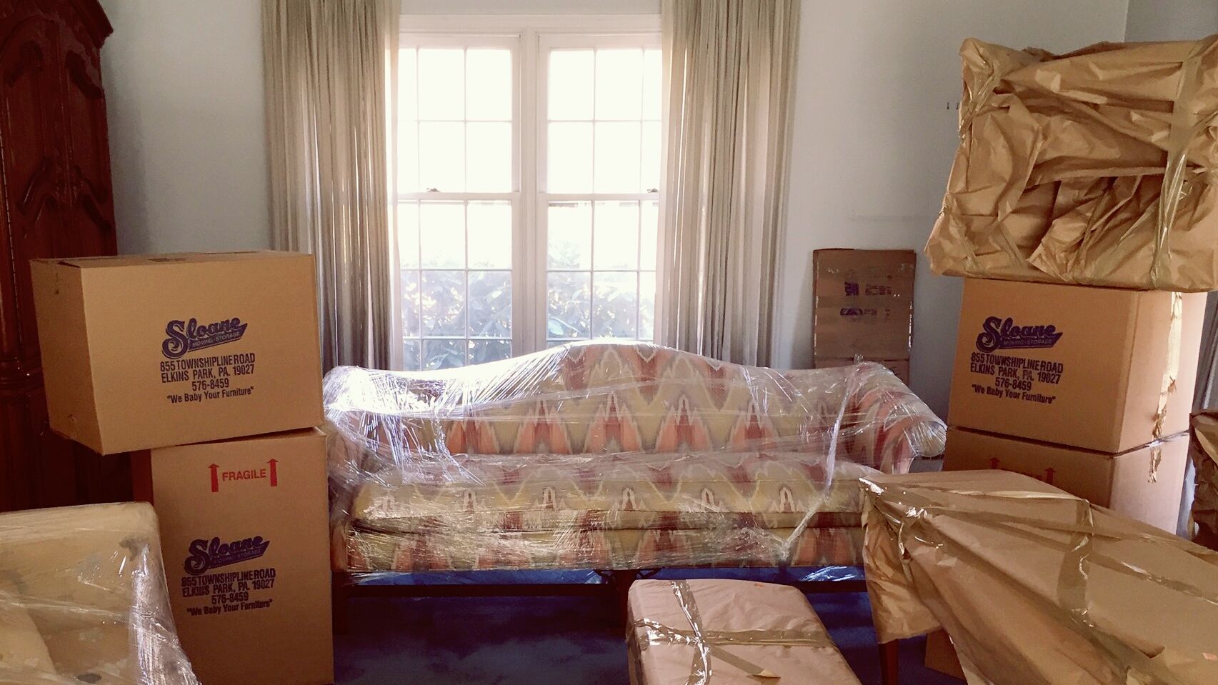Room with furniture wrapped for moving, surrounded by cardboard boxes. Window in background.