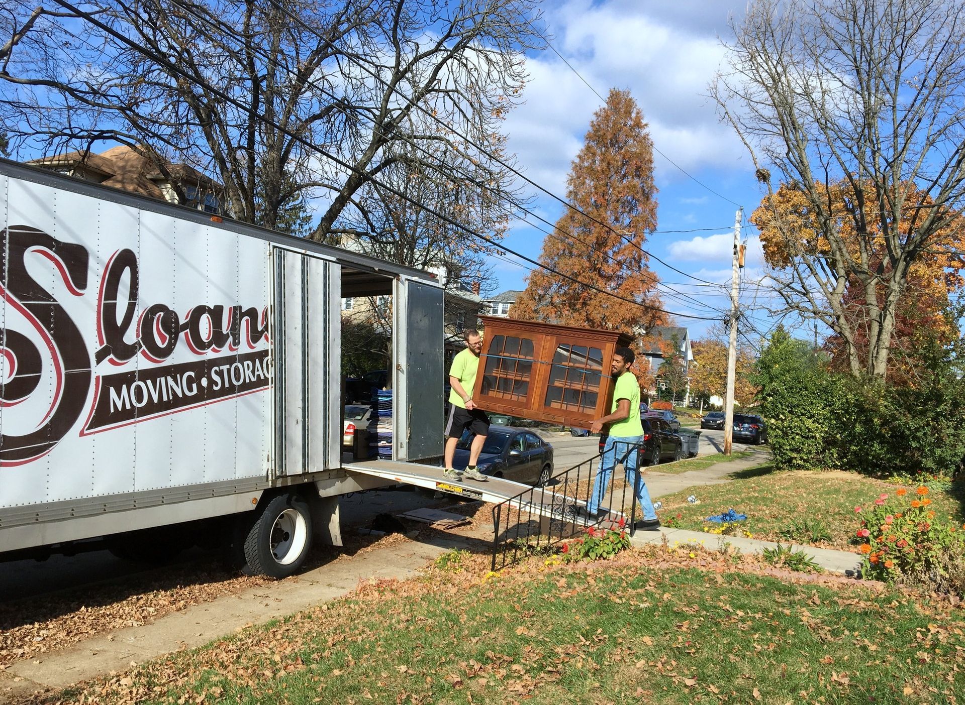 Two movers unload a large wooden cabinet from a Sloan Moving Storage truck on a sunny day.
