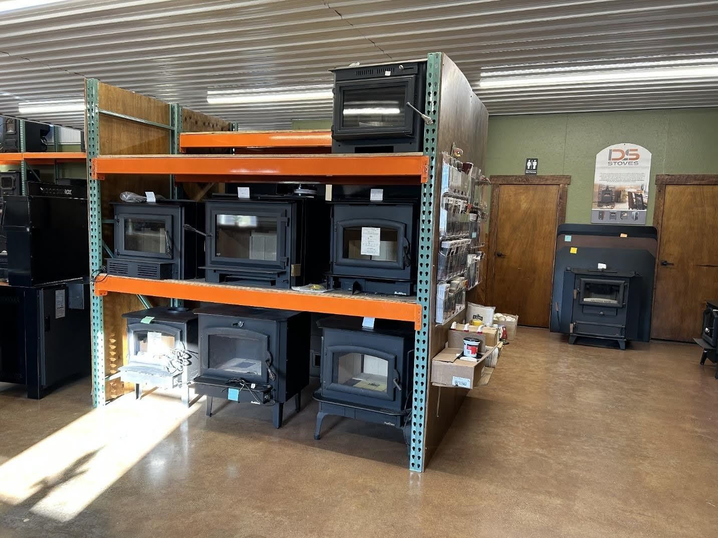 A retail warehouse store displaying various black wood-burning stoves on metal industrial shelving and the floor.
