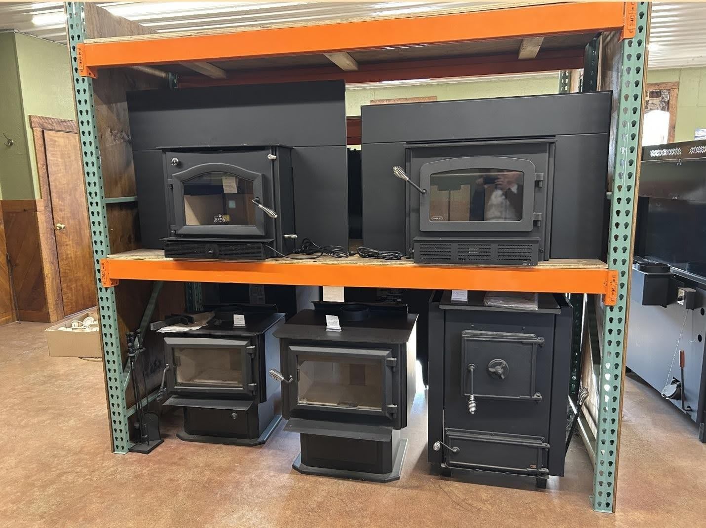 Black wood stoves and fireplace inserts displayed on a metal industrial storage rack in a retail shop.