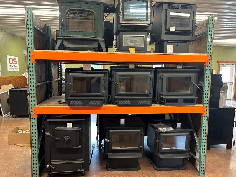Shelves stacked with various black wood-burning stoves in a store; mostly rectangular with glass fronts.