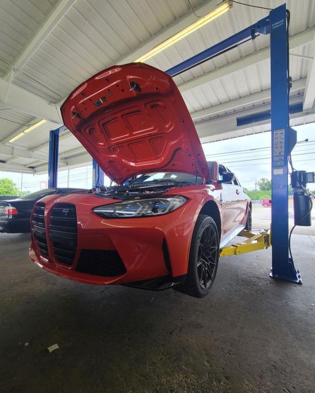 A red car with the hood up is on a lift in a garage.