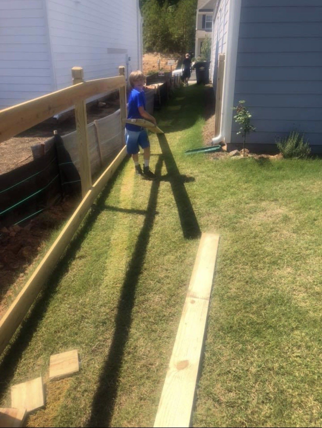 A person carries wood along a grassy yard between two buildings. The unfinished fence is on the left.