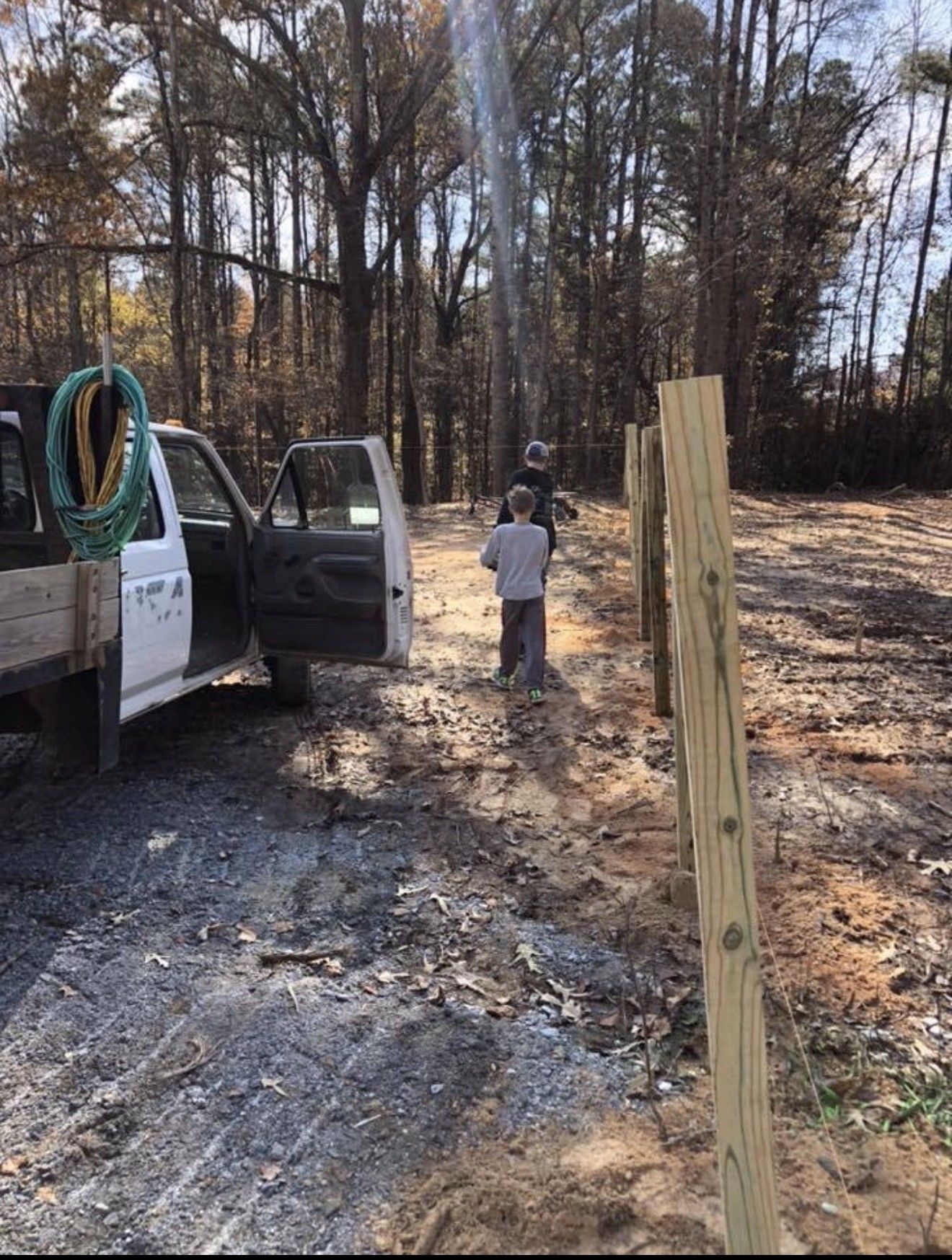 A person and child near a truck and fence posts in a wooded area. The sun is shining.