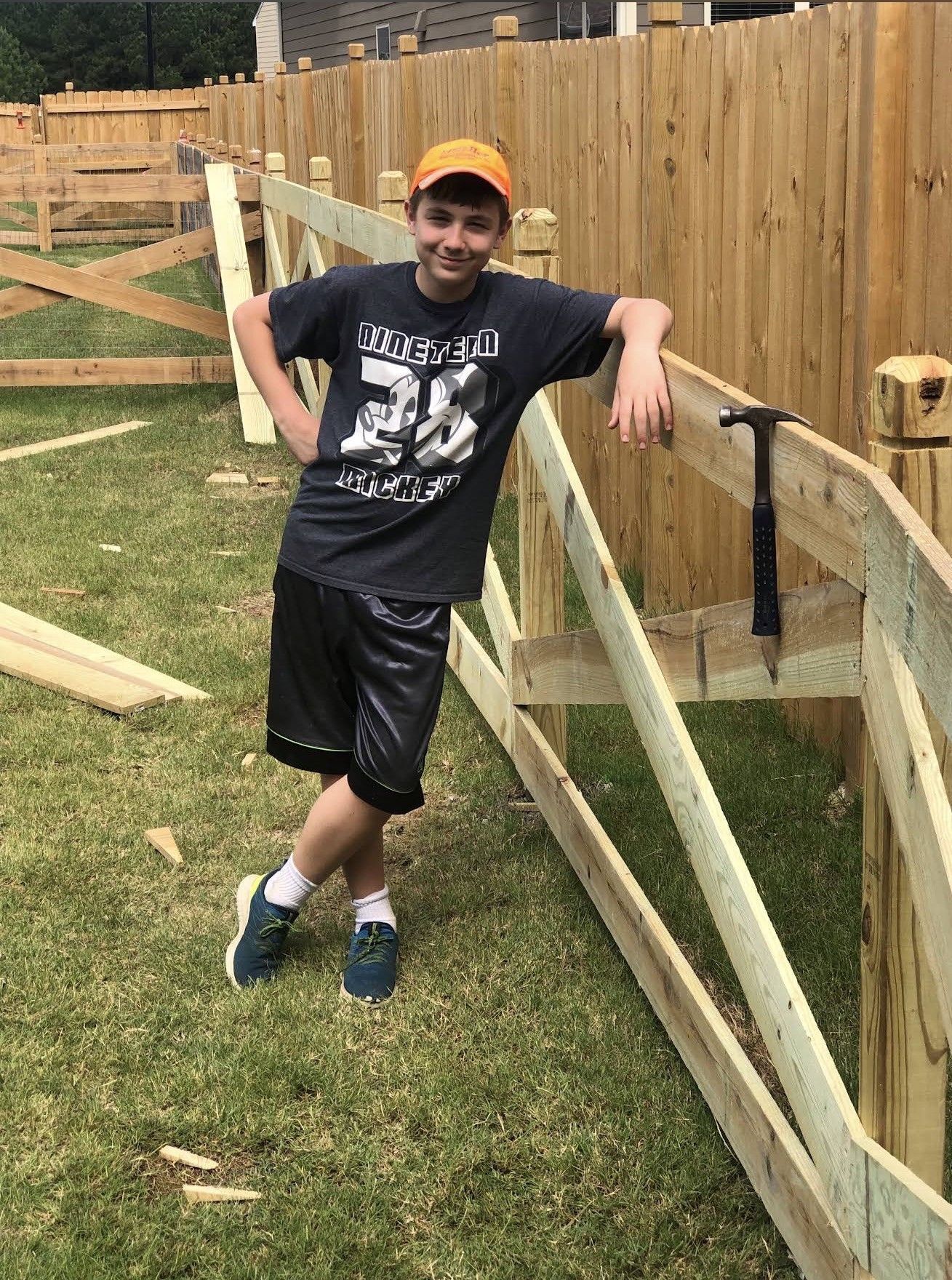 Teenager leans on a partially built wooden fence in a grassy yard, wearing a t-shirt, shorts, and orange hat.