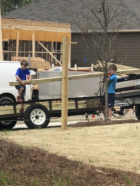 Two children near a trailer at a construction site; one sits, the other stands next to wood planks.