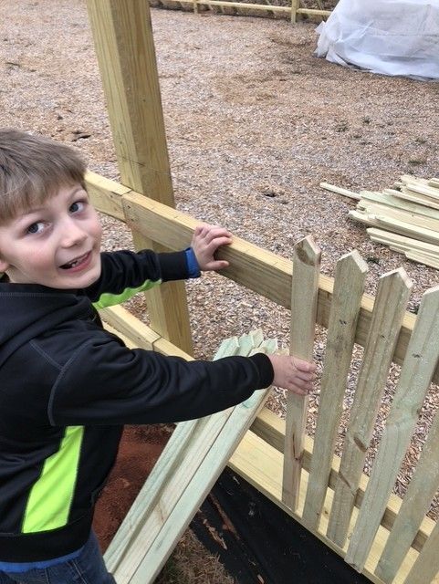 Boy smiles, holding picket fence plank. Wooden fence being built outdoors.