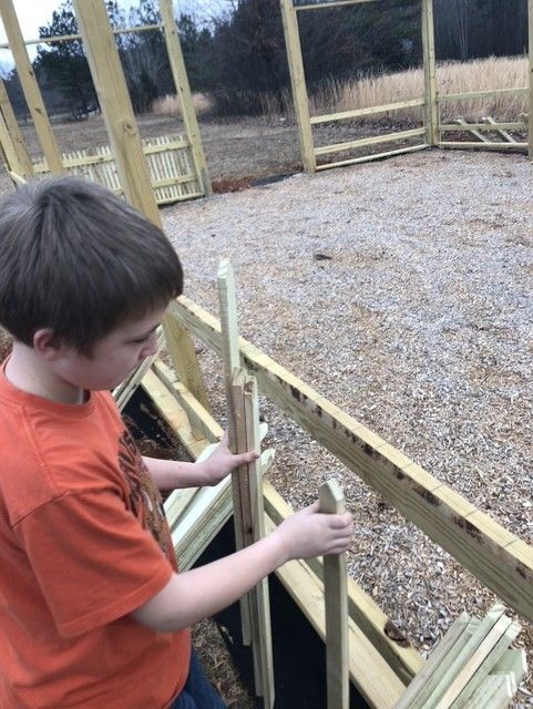 Boy assembling wooden fence. He's outside, near other fence sections. He holds a vertical piece.