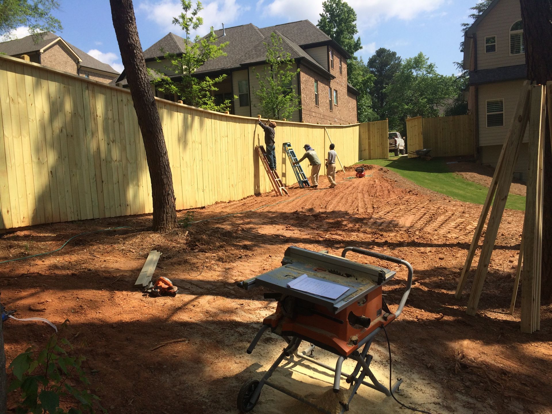 Construction workers building a wooden fence in a backyard, with a saw table in the foreground and houses in the background.