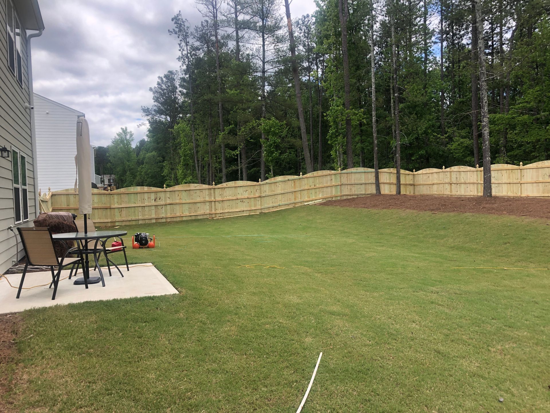 Backyard with new wooden fence, patio furniture, and lawn. Trees in background.