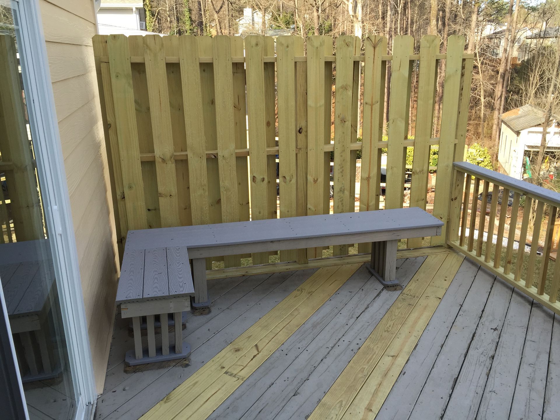 Wooden deck with built-in gray bench. Yellow and gray striped flooring, wooden fence. Sunny outdoor setting.