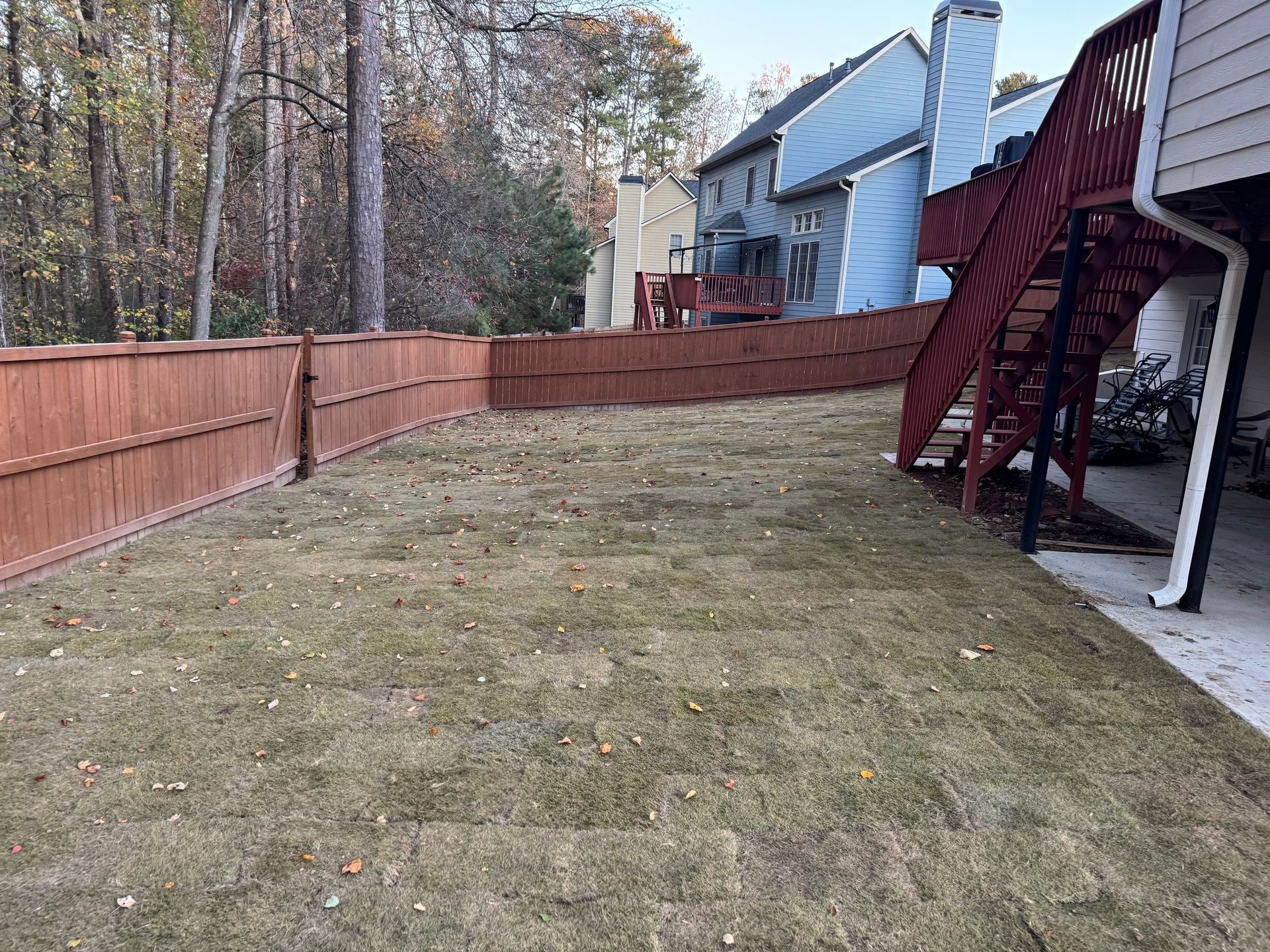 A brown wooden fence surrounds a patchy yard, next to a house with a deck and stairs.