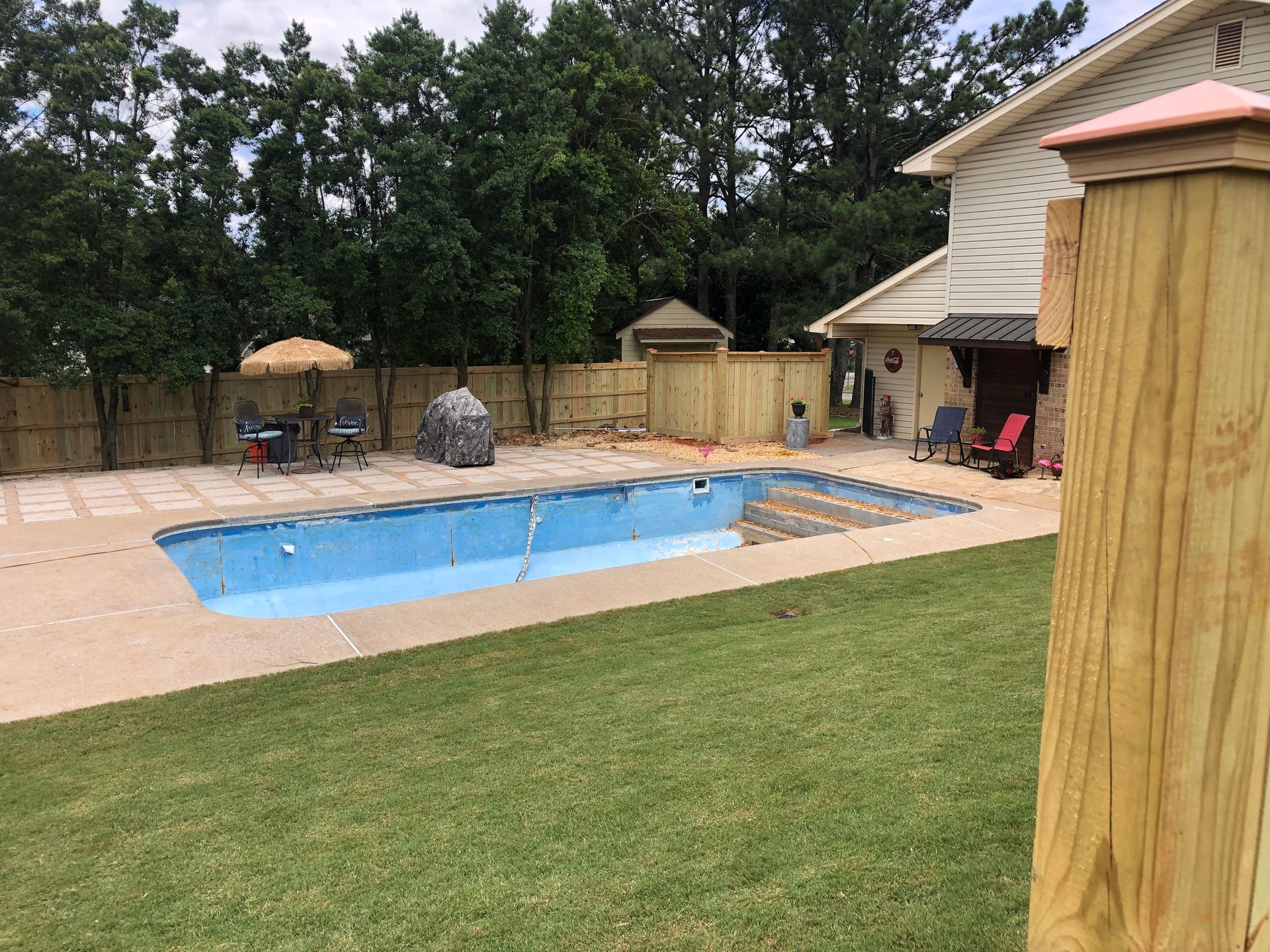 Empty blue swimming pool in backyard with green grass, brown fencing, and a house.