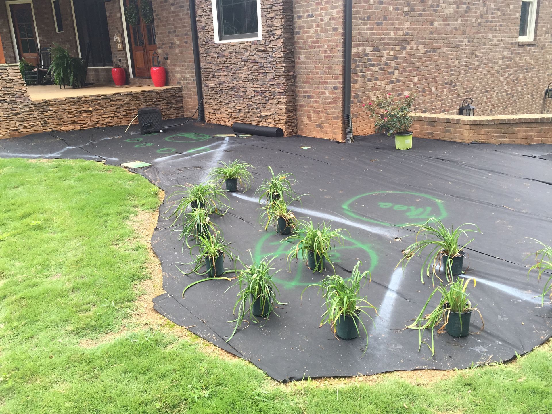 Black landscaping fabric with plants, marked planting areas, and a brick building.