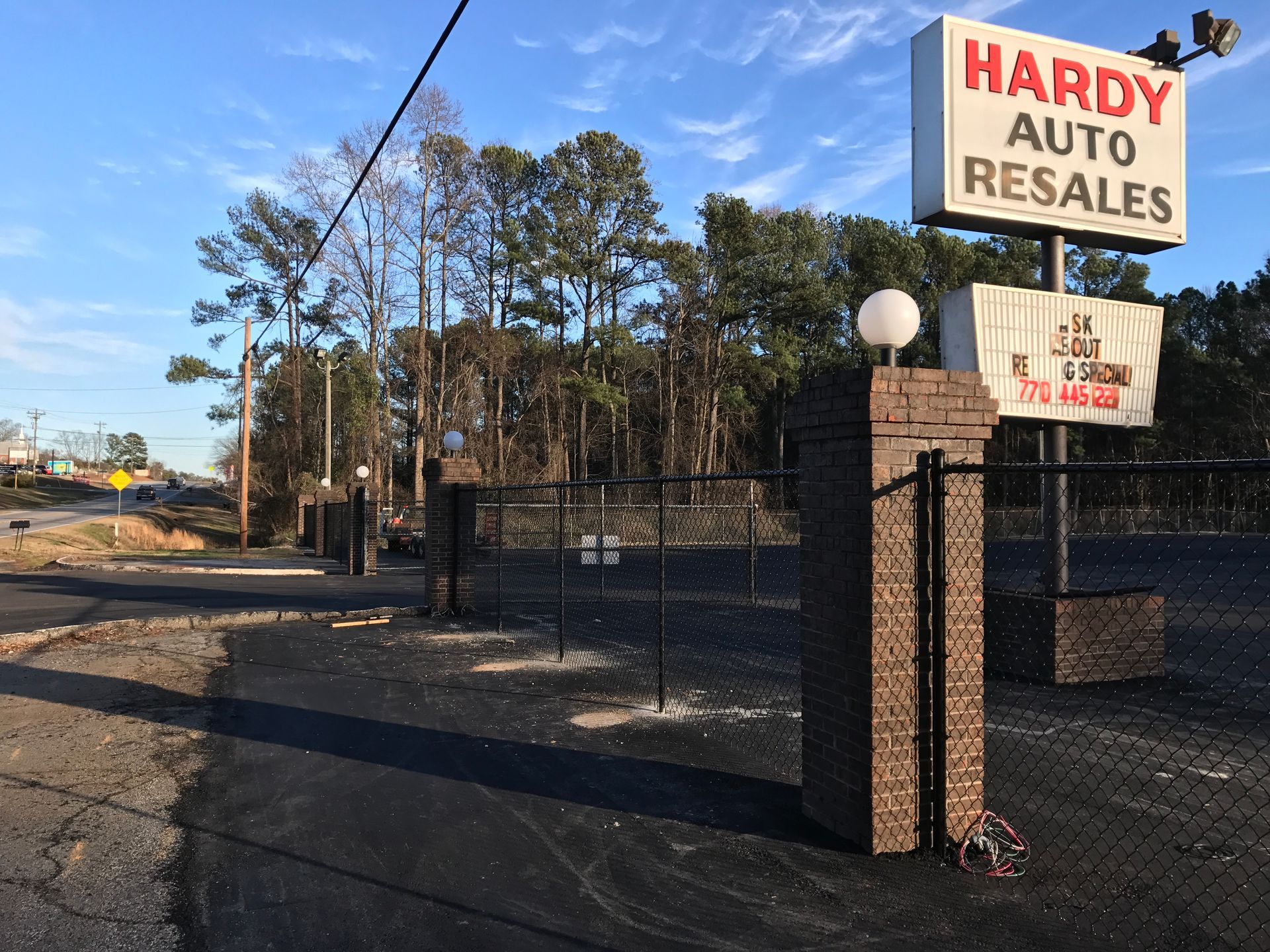 Hardy Auto Resales sign above a fenced lot. Dark pavement and trees in the background under a blue sky.