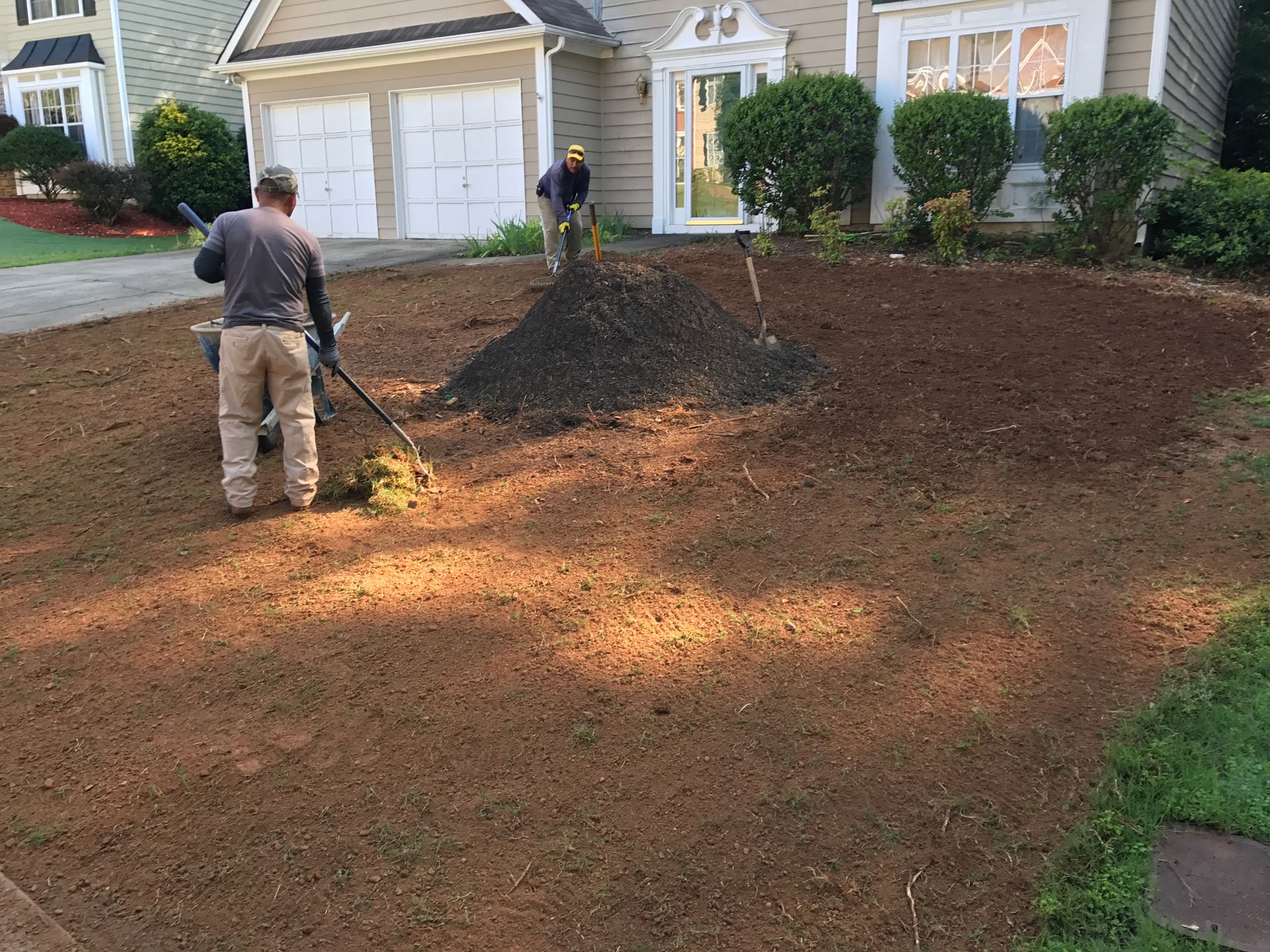 Two people spreading soil in a front yard. One uses a machine, the other a rake near a house.